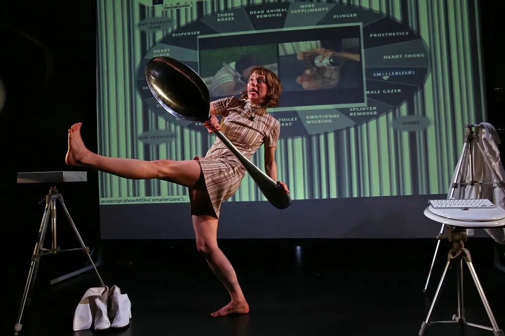 Michelle Ellsworth on stage
holding a person-sized spoon
in front of a projection
of a website
